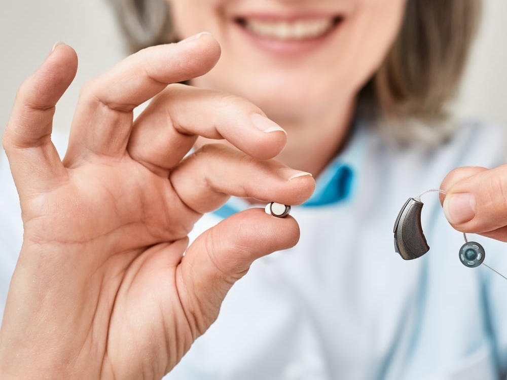 Woman holding a zinc-air hearing aid battery beside her hearing aid, sizes stocked in Falconer, NY